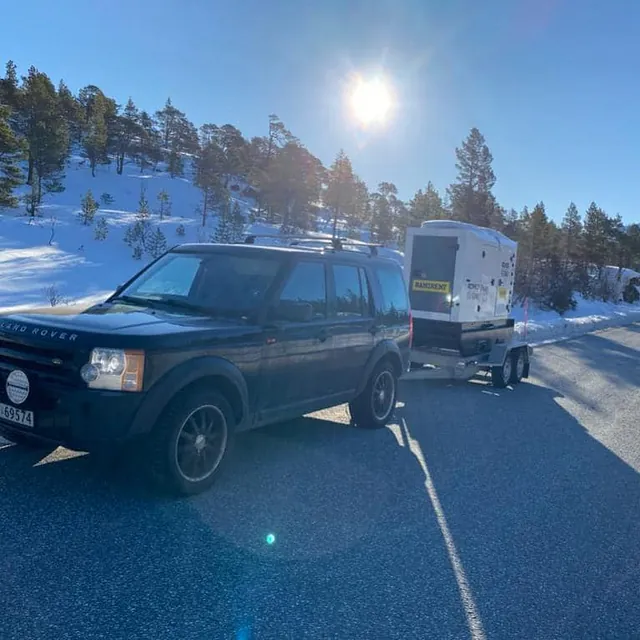 Svart bil med tilhenger og aggregat parkert langs vei i snødekt terreng.