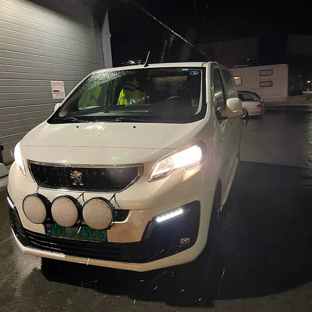 Hvit varebil parkert utenfor verksted om kvelden med lysene på i regnvær.
