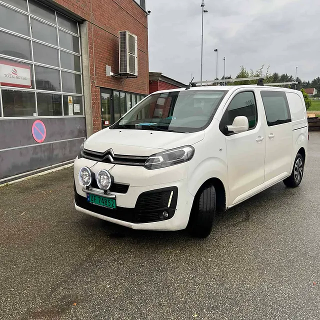 Hvit varebil parkert utenfor industribygg med murfasade og garasjeport.