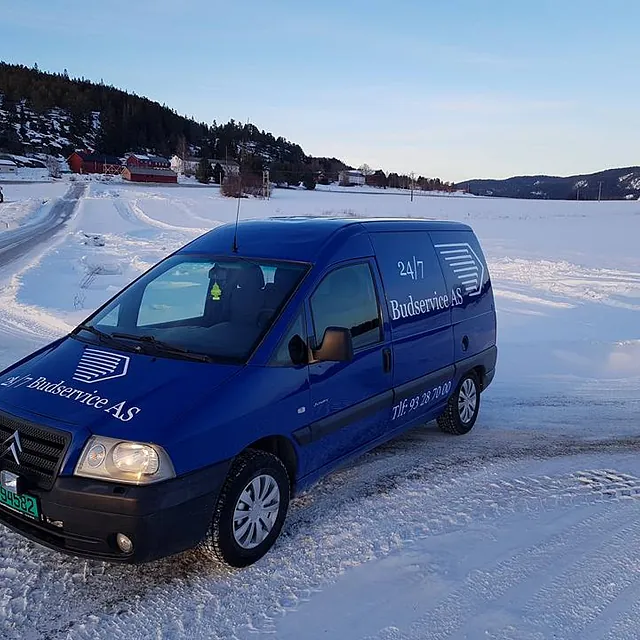 Blå varebil parkert på snødekt jorde med gårdsbygninger i bakgrunnen.