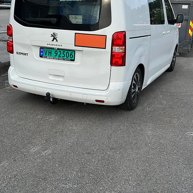 Hvit varebil står parkert på industriområde med containere i bakgrunnen.