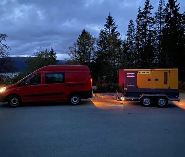 Rød varebil trekker gult aggregat på tilhenger i skumring.
